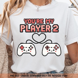 May include: White t-shirt with the text "YOU'RE MY PLAYER 2" in a pixelated font with a pink outline. Below the text are two white game controllers connected by a heart-shaped cable. The shirt is a digital download.