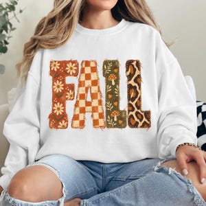 May include: A white crewneck sweatshirt featuring the word FALL printed in large block letters. Each letter has a unique pattern: the F has white daisies on orange, the A is an orange checkerboard, the L has mushrooms on green, and the L is leopard print.