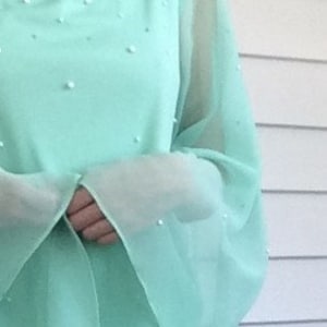 K&ouml;nnte beinhalten: Ein mintgr&uuml;nes Maxikleid mit einem durchsichtigen &Uuml;berwurf und Perlenbesatz. Das Kleid hat einen runden Ausschnitt und lange &Auml;rmel.
