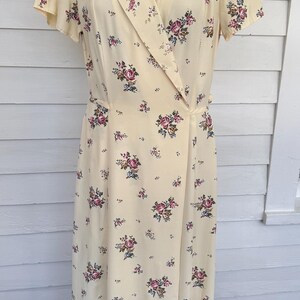 May include: A vintage-style, pale yellow dress with a floral pattern. The dress features a collared neckline, short sleeves, and a wrap-style design. The floral print includes pink, purple, and blue flowers scattered across the fabric. The dress is displayed on a mannequin.