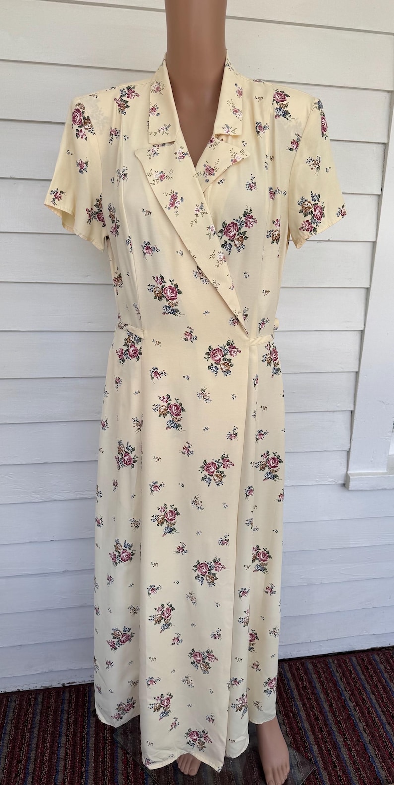 May include: A vintage-style, pale yellow dress with a floral pattern. The dress features a collared neckline, short sleeves, and a wrap-style design. The floral print includes pink, purple, and blue flowers scattered across the fabric. The dress is displayed on a mannequin.