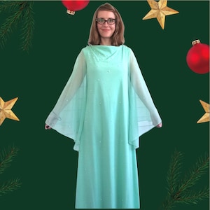 K&ouml;nnte beinhalten: Eine Frau tr&auml;gt ein mintgr&uuml;nes, lang&auml;rmeliges Kleid mit einem Perlenbesatz am Ausschnitt. Das Kleid hat einen flie&szlig;enden, kapenartigen Schnitt und ist perfekt f&uuml;r einen formellen Anlass.