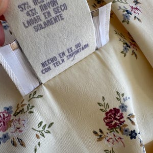 May include: Close-up of a cream-colored fabric with a floral pattern in pink, blue, and burgundy. A tag with text in Spanish reads "57% Acetate, 43% Rayon, Dry Clean Only, Made in the USA, Imported Fabric."