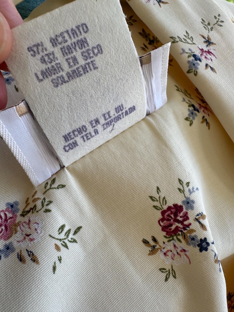 May include: Close-up of a cream-colored fabric with a floral pattern in pink, blue, and burgundy. A tag with text in Spanish reads "57% Acetate, 43% Rayon, Dry Clean Only, Made in the USA, Imported Fabric."