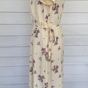 May include: A cream-colored, vintage-style dress with a floral pattern. The dress features short sleeves, a tie waist, and a button-down front. The floral design includes pink, purple, and blue flowers.