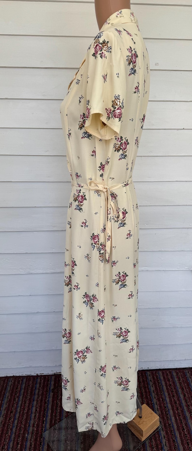 May include: A cream-colored, vintage-style dress with a floral pattern. The dress features short sleeves, a tie waist, and a button-down front. The floral design includes pink, purple, and blue flowers.