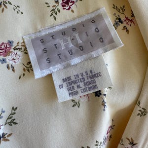 May include: Close-up of a cream-colored fabric with a floral pattern of burgundy, blue, and green. The fabric has a tag that reads "STUDIO THI STUDIO" and "MADE IN U.S.A. OF IMPORTED FABRIC".