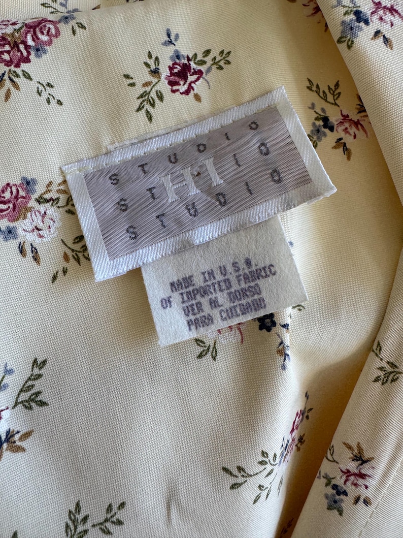 May include: Close-up of a cream-colored fabric with a floral pattern of burgundy, blue, and green. The fabric has a tag that reads "STUDIO THI STUDIO" and "MADE IN U.S.A. OF IMPORTED FABRIC".