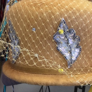 May include: A tan felt hat featuring a yellow netting veil draped over the crown. The hat is decorated with silver sequined leaf-shaped appliqués and small clear rhinestones. A brown grosgrain ribbon band encircles the base of the hat.