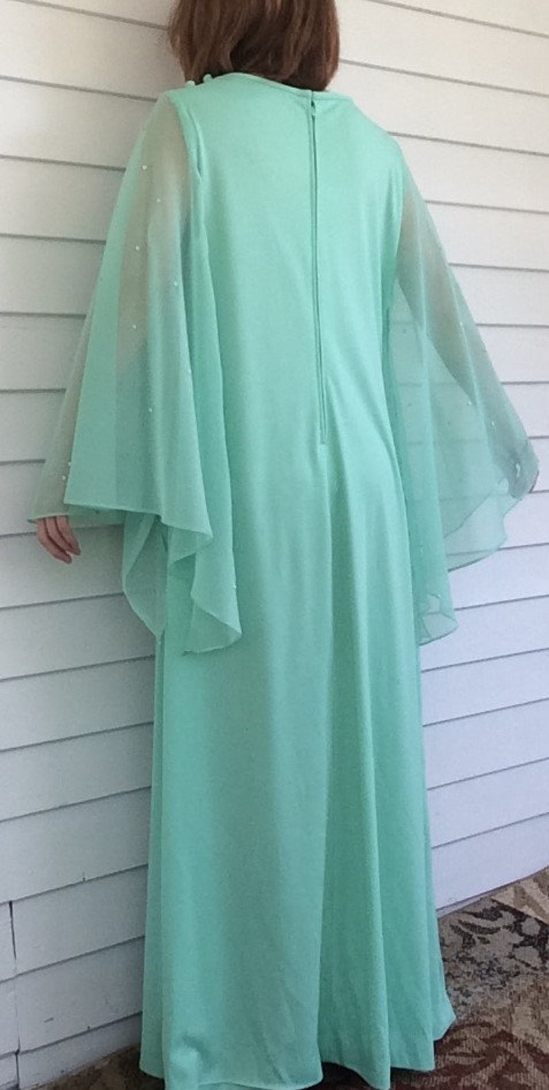 K&ouml;nnte beinhalten: Ein langes, flie&szlig;endes Maxikleid in Mintgr&uuml;n mit durchsichtigen, glockenf&ouml;rmigen &Auml;rmeln. Das Kleid hat einen Rei&szlig;verschluss auf der R&uuml;ckseite und ist aus einem weichen, leichten Stoff gefertigt.