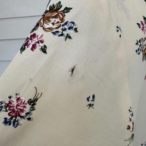May include: Cream-colored fabric with a floral pattern featuring burgundy, brown, and blue flowers. The fabric appears to be part of a garment, possibly a blouse or dress, with a visible sleeve and shoulder.