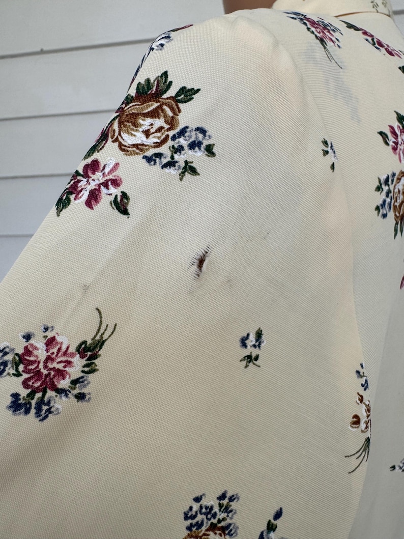 May include: Cream-colored fabric with a floral pattern featuring burgundy, brown, and blue flowers. The fabric appears to be part of a garment, possibly a blouse or dress, with a visible sleeve and shoulder.