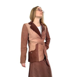 Puede incluir: Una chaqueta de pana marrón con un diseño de patchwork. La chaqueta tiene cuello, mangas largas y cierre de botones. La chaqueta se lleva sobre una camisa blanca y una falda marrón.