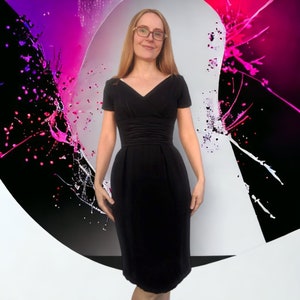 Può includere: Una donna indossa un abito nero al ginocchio con scollo a V e maniche corte. L'abito ha un corpetto aderente e una gonna leggermente svasata. La donna è in piedi davanti a uno sfondo astratto colorato.