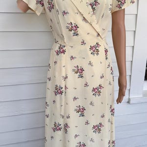 May include: A vintage-style, cream-colored dress with a floral pattern. The dress features a wrap-style bodice with a collar and short sleeves. The fabric is adorned with clusters of pink, purple, and green flowers. The dress is knee-length.