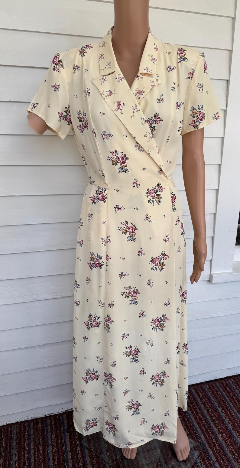 May include: A vintage-style, cream-colored dress with a floral pattern. The dress features a wrap-style bodice with a collar and short sleeves. The fabric is adorned with clusters of pink, purple, and green flowers. The dress is knee-length.