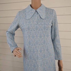Pode incluir: Vestido vintage azul claro com um padrão floral branco. O vestido apresenta mangas compridas e uma gola grande e arredondada. O vestido é exibido em um manequim.