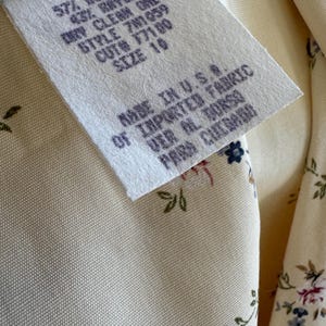 May include: Close-up of a cream-colored fabric with a floral pattern and a tag. The tag reads "RN 50640, 57% Acetate, 43% Rayon, Dry Clean Only, Style 701059, Cut 77100, Size 10, Made in U.S.A. of Imported Fabric."