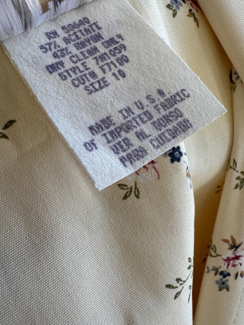 May include: Close-up of a cream-colored fabric with a floral pattern and a tag. The tag reads "RN 50640, 57% Acetate, 43% Rayon, Dry Clean Only, Style 701059, Cut 77100, Size 10, Made in U.S.A. of Imported Fabric."