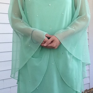 K&ouml;nnte beinhalten: Ein mintgr&uuml;nes Maxikleid mit langen &Auml;rmeln und einem hohen Ausschnitt. Das Kleid ist mit kleinen wei&szlig;en Perlen verziert.