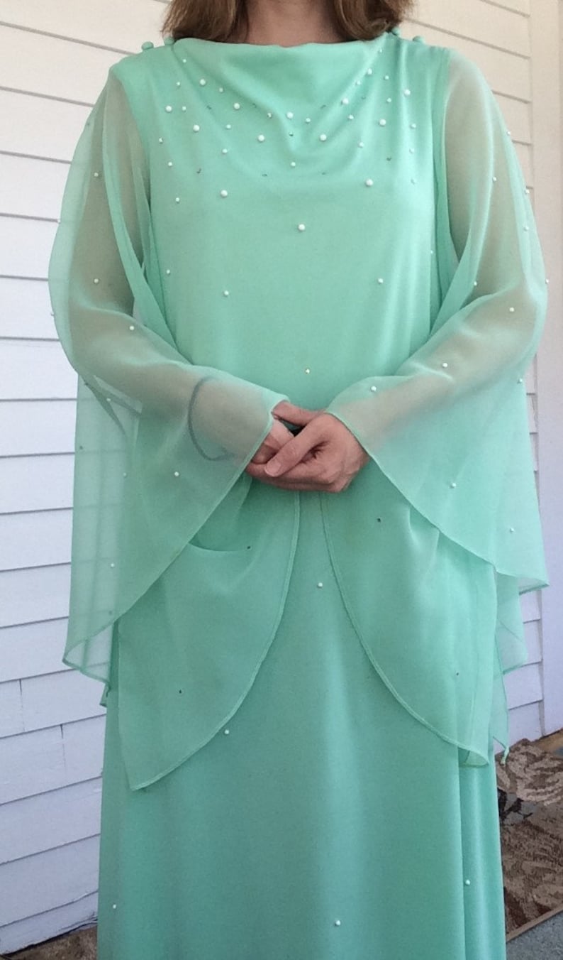 K&ouml;nnte beinhalten: Ein mintgr&uuml;nes Maxikleid mit langen &Auml;rmeln und einem hohen Ausschnitt. Das Kleid ist mit kleinen wei&szlig;en Perlen verziert.