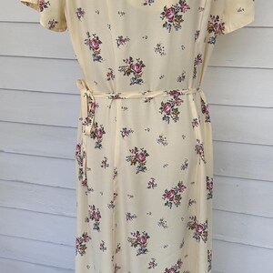 May include: A light yellow dress with a floral pattern of pink and blue roses. The dress has short sleeves and a tie belt at the waist. The dress is approximately 140 cm in length.
