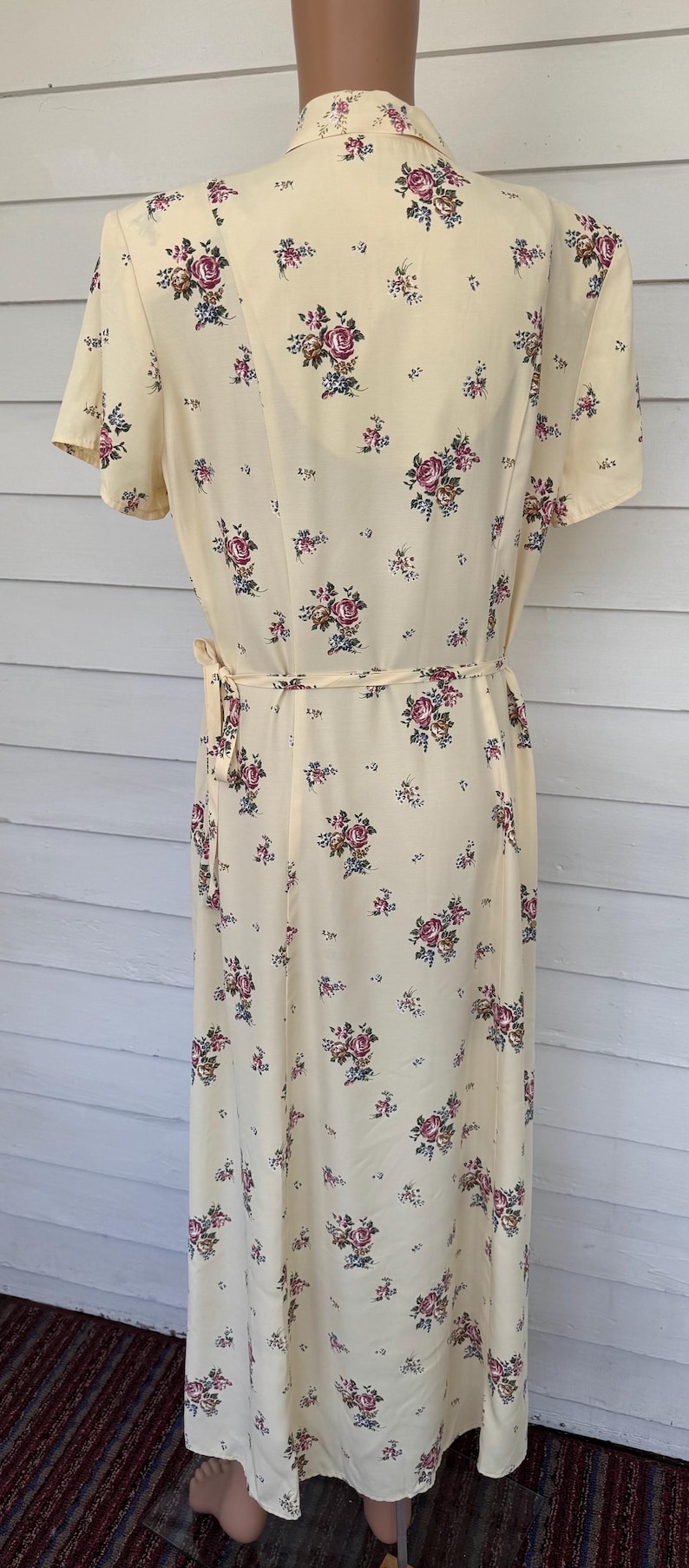 May include: A light yellow dress with a floral pattern of pink and blue roses. The dress has short sleeves and a tie belt at the waist. The dress is approximately 140 cm in length.