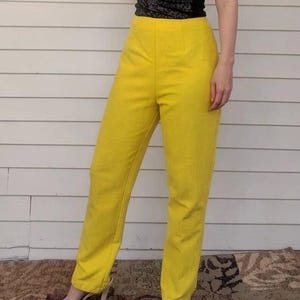 Puede incluir: Pantalones amarillos de cintura alta. Los pantalones tienen un corte a medida con una pernera recta. La tela parece ser un tejido texturizado. Los pantalones se combinan con un top negro. El fondo es una pared blanca.