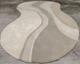 Op de afbeelding: Een onregelmatig gevormd vloerkleed met een vloeiend, organisch ontwerp met gelaagde kleurstroken. Het palet bestaat uit zachte crème, lichtbeige en gedempte saliegroene tinten, gerangschikt in gladde, gebogen, golfachtige patronen over het oppervlak.