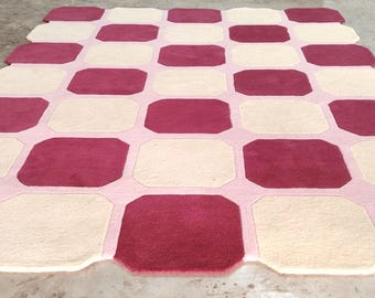 Op de afbeelding: Een groot, geometrisch tapijt met een patroon van afwisselende bordeauxrode en crèmekleurige achthoeken. De achthoeken zijn omlijnd met een lichtroze rand, wat een visueel aantrekkelijk ontwerp creëert. Het tapijt is gemaakt van een zacht, pluche materiaal.