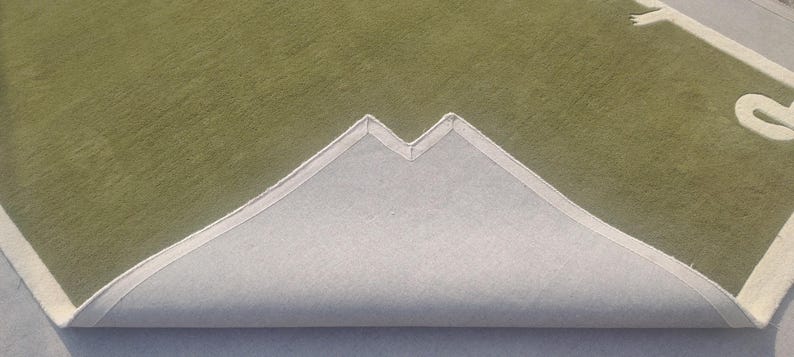 Op de afbeelding: Een groen tapijt met een witte rand en een wit ontwerp in de hoek. Het tapijt is gedeeltelijk teruggevouwen en onthult een lichtgrijze achterkant.