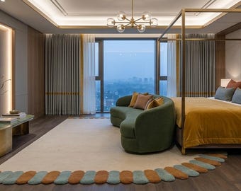 Op de afbeelding: Een luxe slaapkamer met een hemelbed, een mosterdkleurig deken en decoratieve kussens. Een groene gebogen bank staat voor een groot raam. Een patroonkleed en een gouden salontafel maken de elegante ruimte compleet.