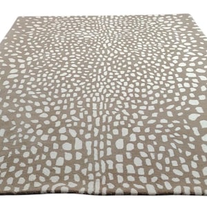 May include: A rectangular area rug with a taupe background and a pattern of white, irregular shapes. The rug's design features a dense, speckled appearance, creating a textured visual effect. The rug is suitable for living rooms and bedrooms.