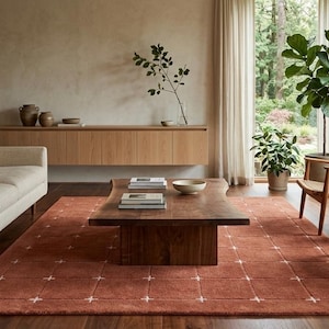 Puede incluir: Un salón moderno con un sofá beige, una mesa de centro de madera oscura y dos sillas de madera con asientos tejidos. Una alfombra de color óxido con un patrón geométrico cubre el suelo. Grandes ventanales ofrecen vistas al exterior.