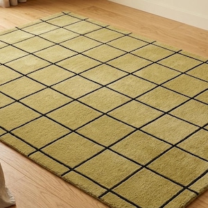 Pode incluir: Um tapete retangular verde-oliva com um padrão de grade de linhas pretas. O tapete está colocado sobre um chão de madeira. Uma pequena mesa de madeira com uma caneca e um cesto de vime com uma manta são visíveis em primeiro plano.