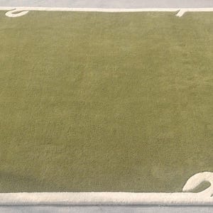 Op de afbeelding: Een rechthoekig groen tapijt met een cr&egrave;mekleurige rand en decoratieve hoekaccenten. Het tapijt heeft een zachte, pluche textuur en een eenvoudig, elegant ontwerp. Afmetingen: ongeveer 244 x 305 cm.