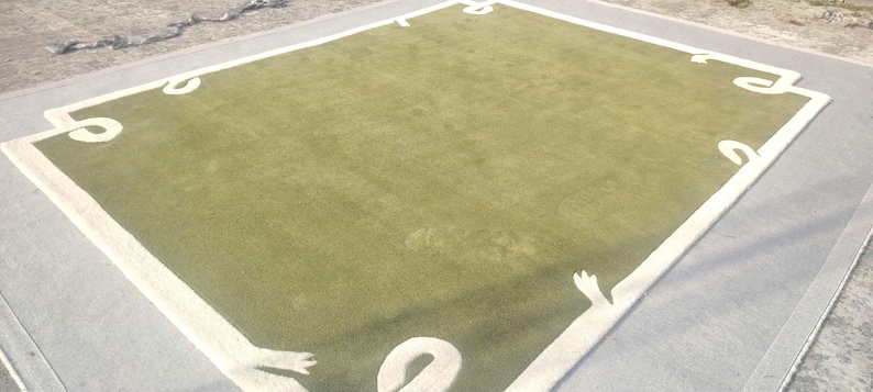Op de afbeelding: Een groot, rechthoekig tapijt met een lichtgroen midden en een witte rand. De rand heeft decoratieve witte vormen in elke hoek. Het tapijt ligt op een grijze ondergrond, waardoor een gelaagd effect ontstaat. Het ontwerp van het tapijt voegt een vleugje elegantie toe.