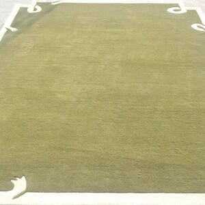 Op de afbeelding: Een rechthoekig groen tapijt met een witte rand en decoratieve hoekaccenten. Het tapijt heeft een effen groen midden en een witte rand met gestileerde, gebogen ontwerpen in elke hoek. Het tapijt is waarschijnlijk gemaakt van zacht, geweven materiaal.