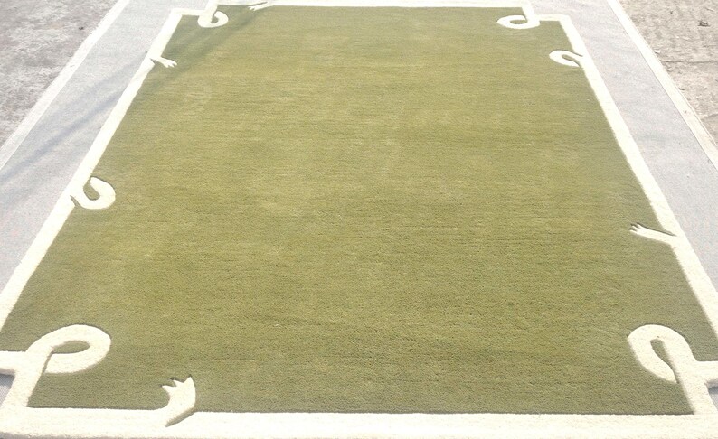 Op de afbeelding: Een rechthoekig groen tapijt met een witte rand en decoratieve hoekaccenten. Het tapijt heeft een effen groen midden en een witte rand met gestileerde, gebogen ontwerpen in elke hoek. Het tapijt is waarschijnlijk gemaakt van zacht, geweven materiaal.