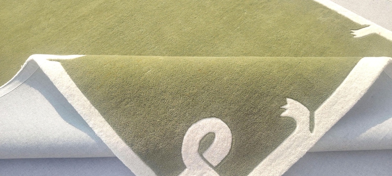 Op de afbeelding: Een opgevouwen groene vloerkleed met een witte rand en een gestileerd ontwerp. Het ontwerp bevat een wit ampersand-symbool en een vogelomtrek. Het vloerkleed heeft een zacht, getextureerd uiterlijk.
