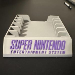 May include: A white Super Nintendo Entertainment System cartridge holder. The holder has a rectangular base with the words "SUPER NINTENDO ENTERTAINMENT SYSTEM" in purple. It features multiple slots for game cartridges.