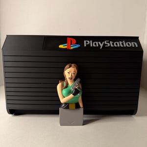 May include: A black PlayStation console replica with the word "PlayStation" printed on the front. A figurine of a woman in a green top and holding a gun is in front of the console. The PlayStation logo is in the upper right corner.