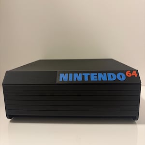 May include: A black Nintendo 64 console with the brand name in blue and the number 64 in red. The console has a rectangular shape with horizontal lines across the front.