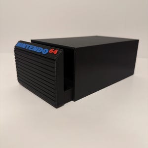 Peut inclure: Une boîte de rangement rectangulaire noire avec une façade de tiroir coulissant présentant des rainures horizontales. Le panneau avant affiche le texte bleu et rouge NINTENDO 64. L'unité a une finition mate lisse et un espace intérieur creux.