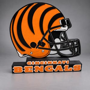 May include: A decorative Cincinnati Bengals football helmet sign featuring a bright orange shell with black tiger stripes and a black face mask. The helmet sits on a black rectangular base with the text Cincinnati Bengals in orange letters.