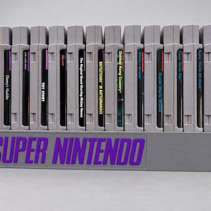Puede incluir: Un soporte de almacenamiento de plástico gris que sostiene trece cartuchos de videojuegos Super Nintendo en fila. La parte frontal del soporte presenta el texto SUPER NINTENDO en letras moradas en negrita. Los cartuchos están dispuestos verticalmente.