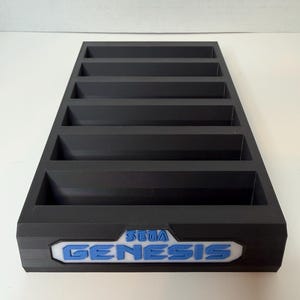 May include: A black rectangular cartridge organiser with multiple slots. The front features the "SEGA GENESIS" logo in blue and white. The organiser is designed to hold and display video game cartridges.