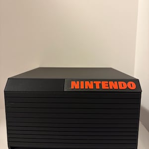 May include: A black console with horizontal lines and the word "NINTENDO" in orange letters. The console has a rectangular shape and is sitting on a white surface.