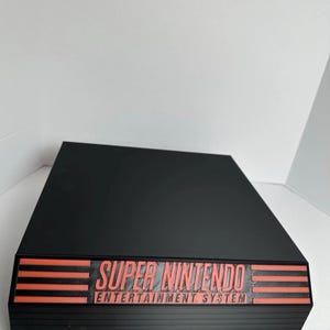 May include: A black Super Nintendo Entertainment System console with red lettering. The console is rectangular with horizontal lines on the front and the words "SUPER NINTENDO ENTERTAINMENT SYSTEM".