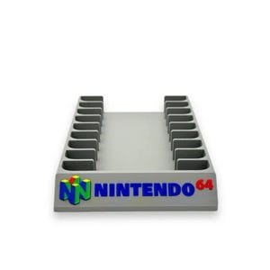 May include: A light grey plastic storage rack for video game cartridges featuring the Nintendo 64 logo on the front. The unit has two parallel rows of vertical slots designed to hold multiple game cartridges in an upright position for organisation.